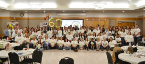 A large group of people in white t-shirts posing together with 60th anniversary balloons.