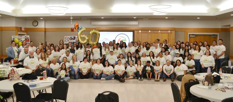 A large group of people in white t-shirts posing together with 60th anniversary balloons.