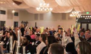 People seated at tables inside a hall during the 2026 Annual Gala, with a woman raising her hand holding a glowing item.