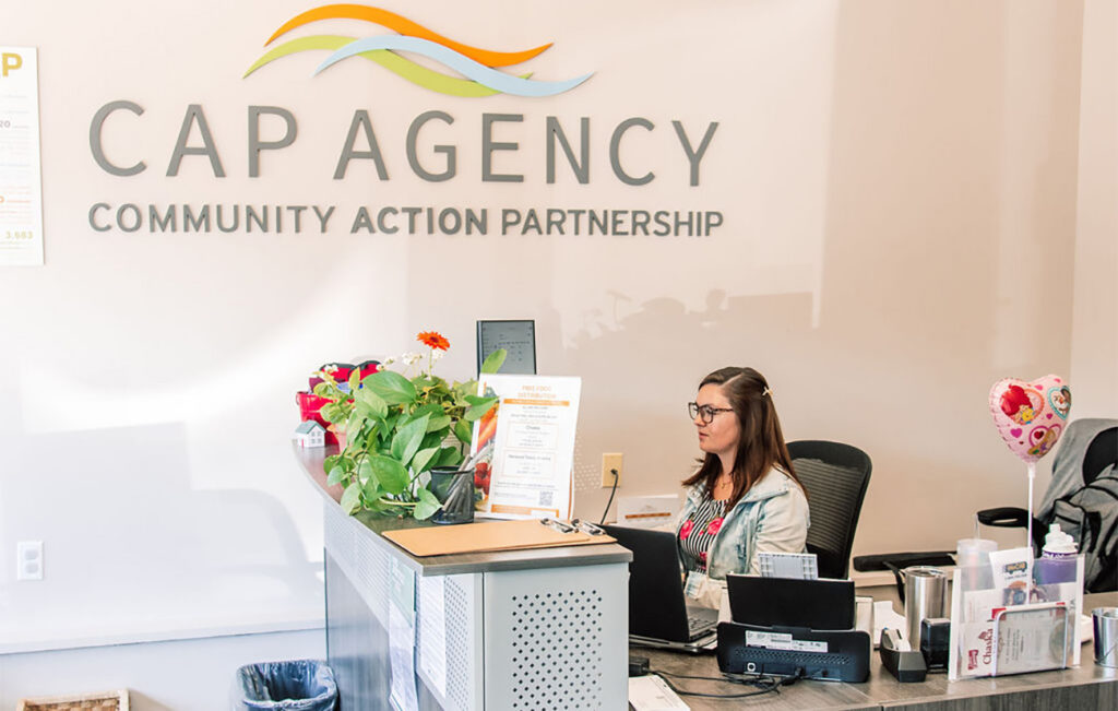 A woman working at the front reception desk inside the CAP Agency office, with the large CAP Agency Community Action Partnership logo displayed on the wall behind her.