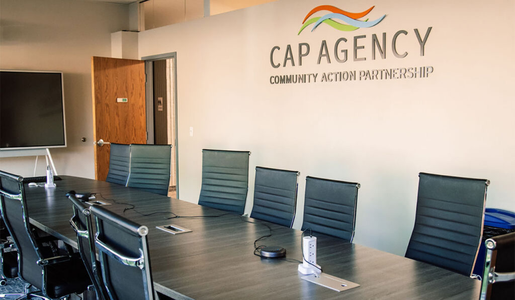 A brightly lit conference room featuring a long meeting table with black office chairs, and the CAP Agency Community Action Partnership logo displayed prominently on the wall.