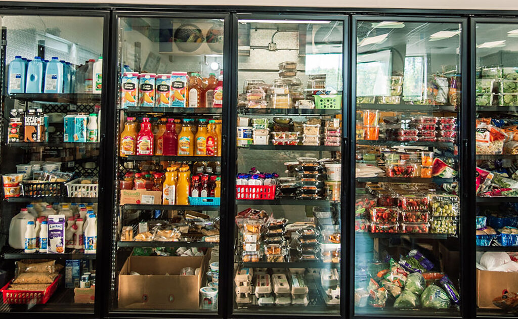 A large commercial refrigerated glass display case in a food pantry filled with shelves of milk, juice bottles, and various packaged food items.