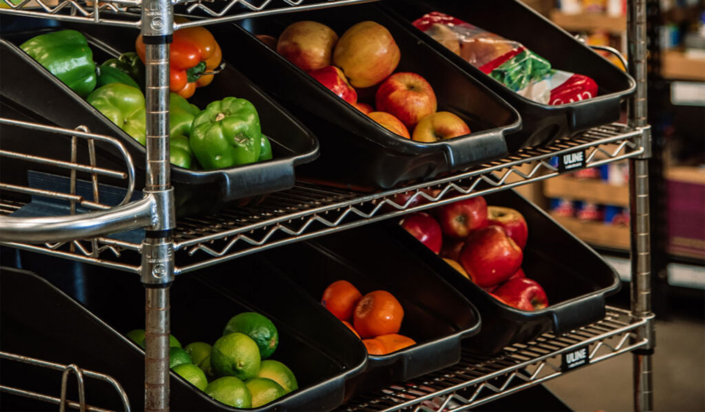 Shelves in a food shelf pantry stocked with fresh apples, green peppers, and limes in black plastic bins.