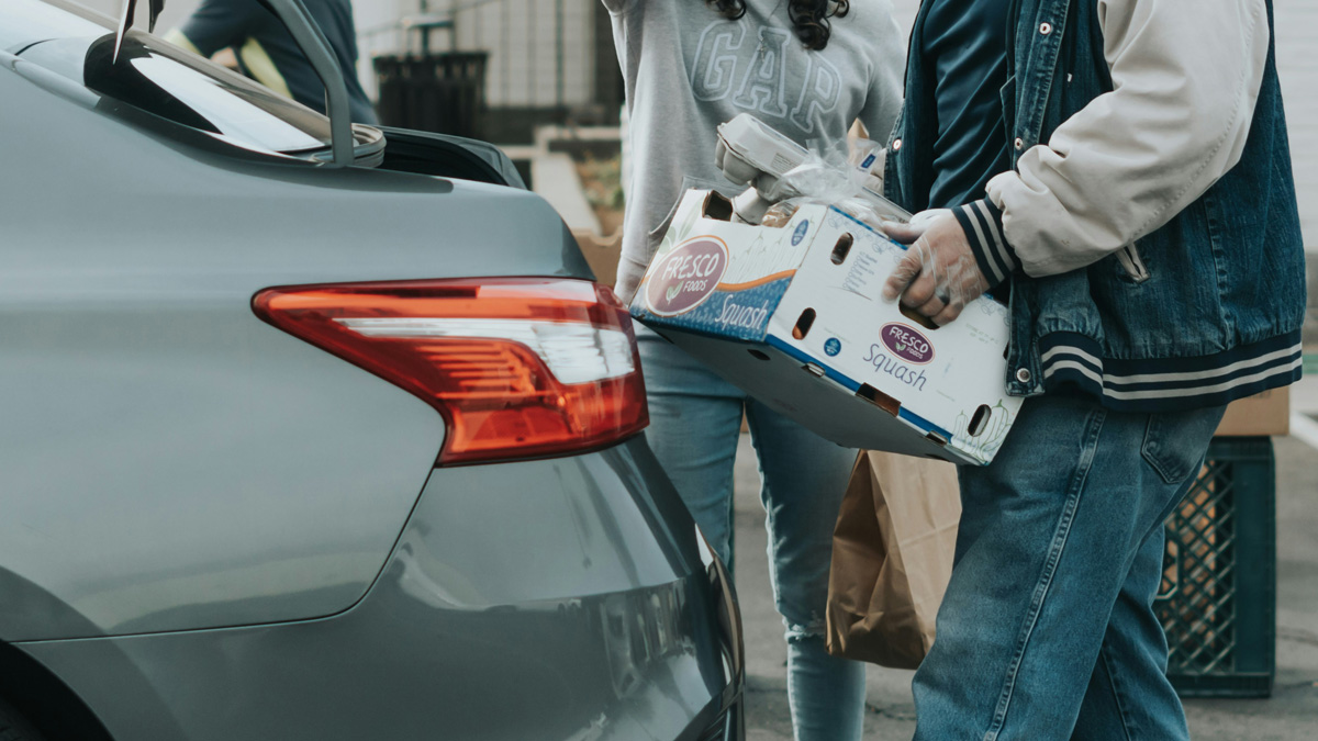 A person carrying a box of food and a brown paper bag toward the open trunk of a silver car