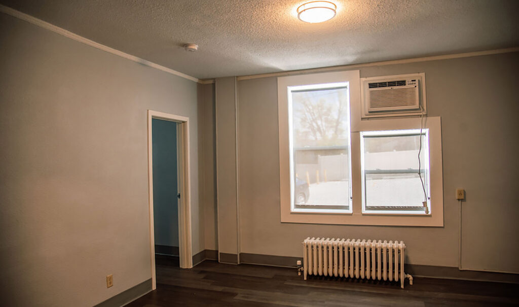 An interior room with light grey walls, dark wood-style flooring, a white-framed window with an air conditioning unit, and a white radiator below.