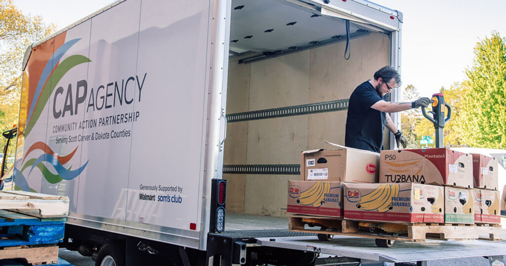 A CAP Agency staff member using a pallet jack to unload boxes of fresh produce, including bananas and plantains, from the back of a delivery truck.