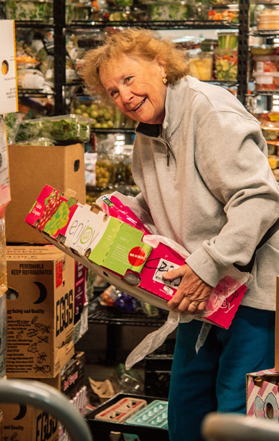 A smiling older female volunteer carrying a box of food through a stocked pantry aisle at the CAP Agency food shelf.