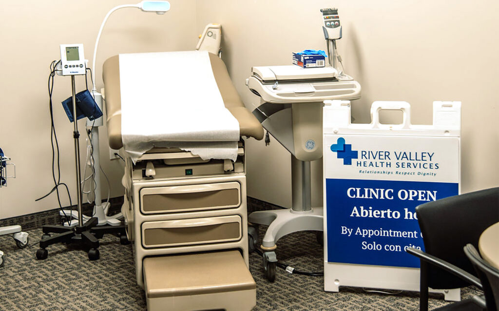 Medical examination room with an exam table, equipment, and a sign for River Valley Health Services Clinic.