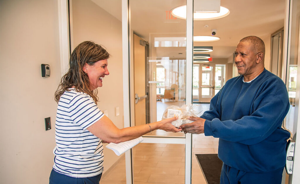 A smiling woman handing a package of food to a senior man indoors.