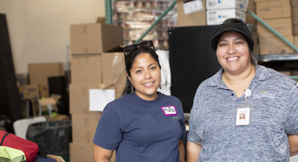 wo smiling volunteers standing in the CAP Agency food shelf warehouse, surrounded by stacks of cardboard boxes.
