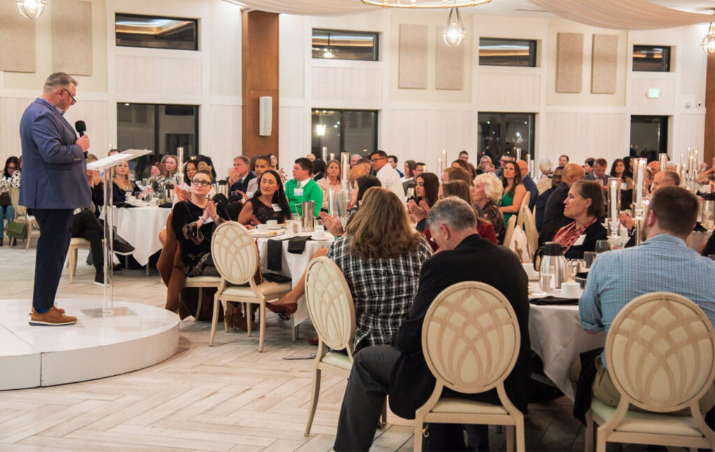A speaker stands at a podium addressing a large audience seated at round tables during a gala event.