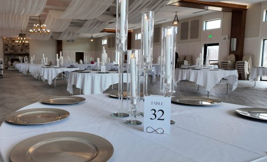 A formal event dining table setup with white linens, silver charger plates, and tall glass candle centerpieces next to a table number 32 card.