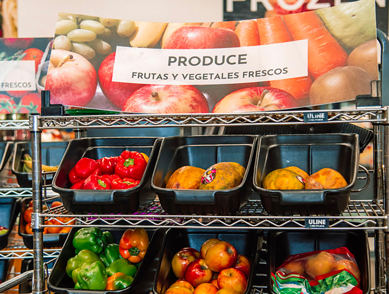 metal display rack in a food pantry containing black bins filled with fresh apples, red bell peppers, and other produce, with a sign above that reads "PRODUCE" and "FRUTAS Y VEGETALES FRESCOS."