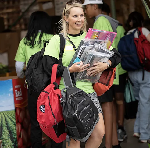 A smiling volunteer wearing a green t-shirt holding several backpacks at an outdoor community event.