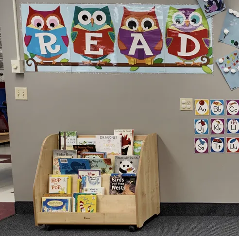 A wooden classroom shelf displaying children's books, storage bins, and colorful blocks spelling the word "READ".